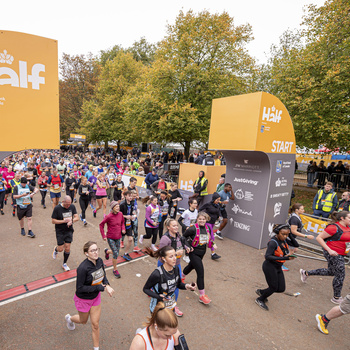 Participants start running in the Royal Parks Half Marathon, passing under arches labeled with the event's name flanked by sponsor signs, in a park setting with autumn foliage.